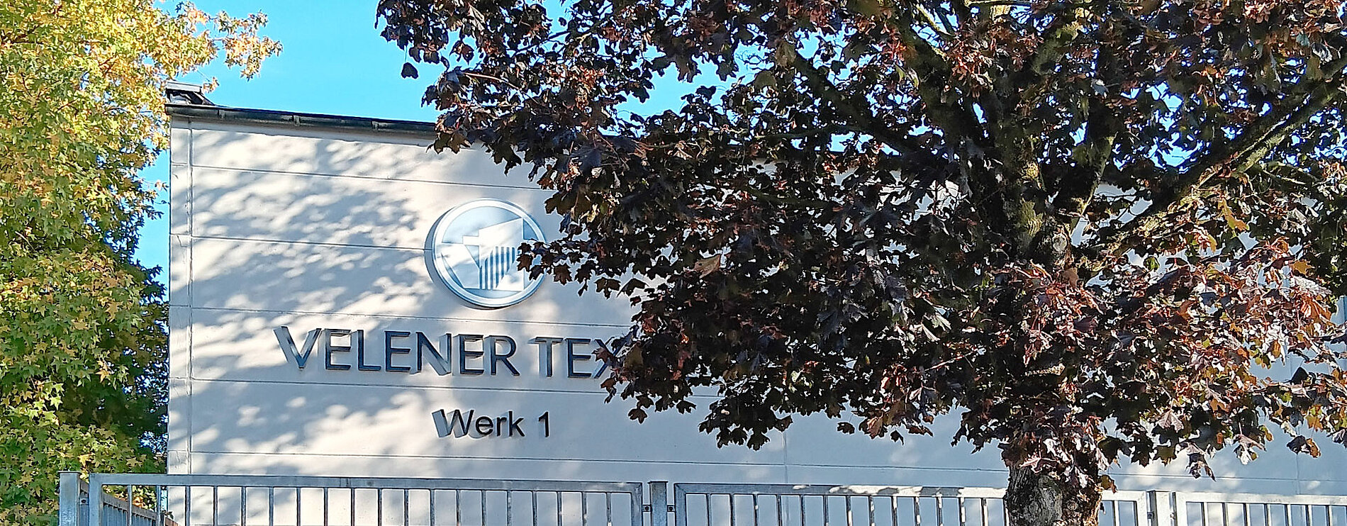 Gebäude von Velener Textil mit dem Schriftzug „Velener Textil – Werk 1“, umgeben von Bäumen